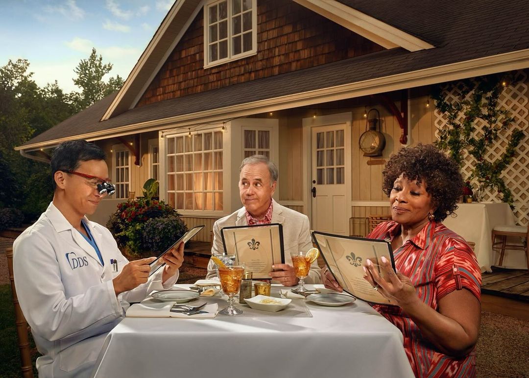Three people seated at a table outside a house, reading menus, with one person in a lab coat and eyeglasses, dining setting, daytime.